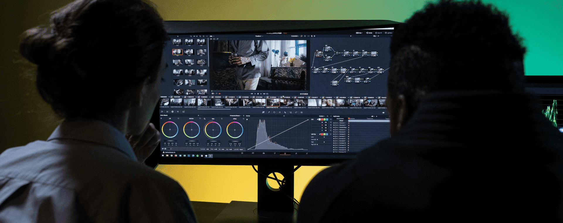Two people sit in front of a computer monitor working on video editing software. The screen displays a video timeline, controls, and color grading tools. The room is dimly lit, with a green and yellow gradient background, setting the stage for a B2B marketing project.