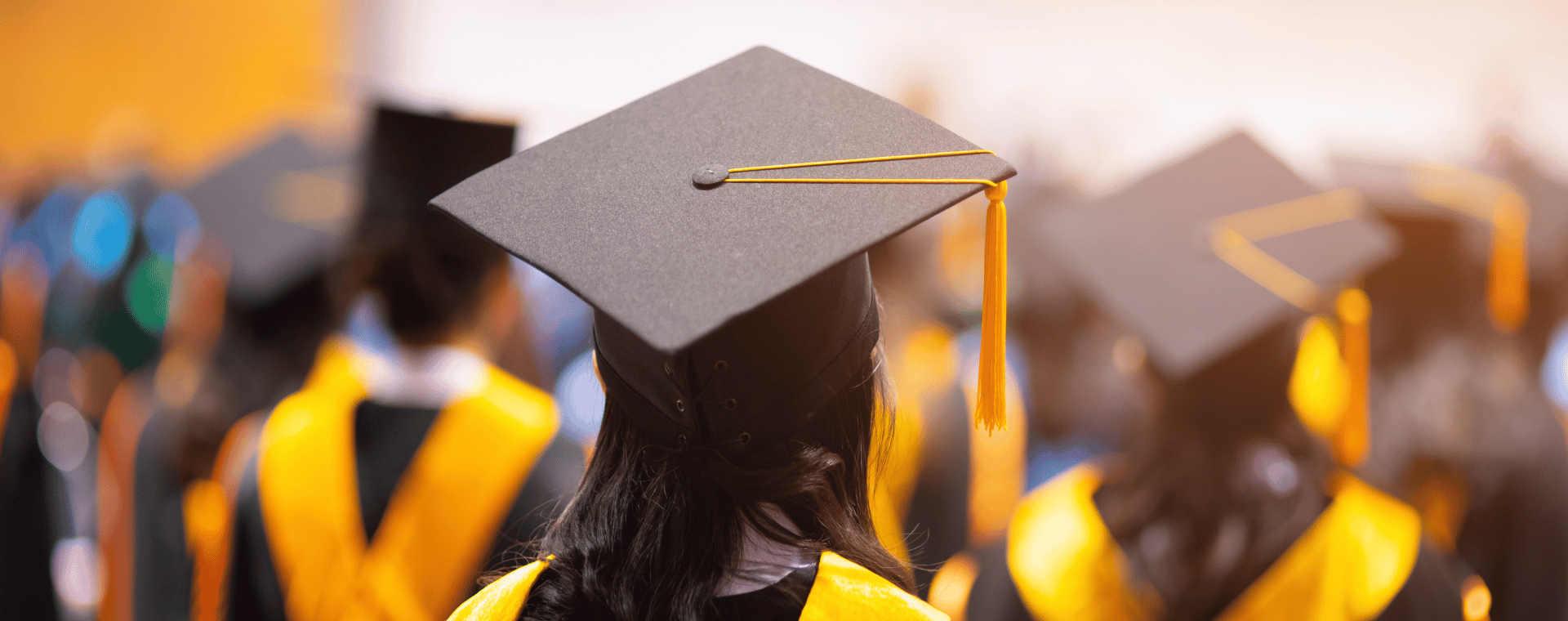 A group of graduates in caps and gowns stands together, facing away from the camera. They are wearing black academic caps with yellow tassels and black gowns with yellow stoles. The background is blurred, creating a focus on the graduates, perfect for use in academic videos highlighting success.