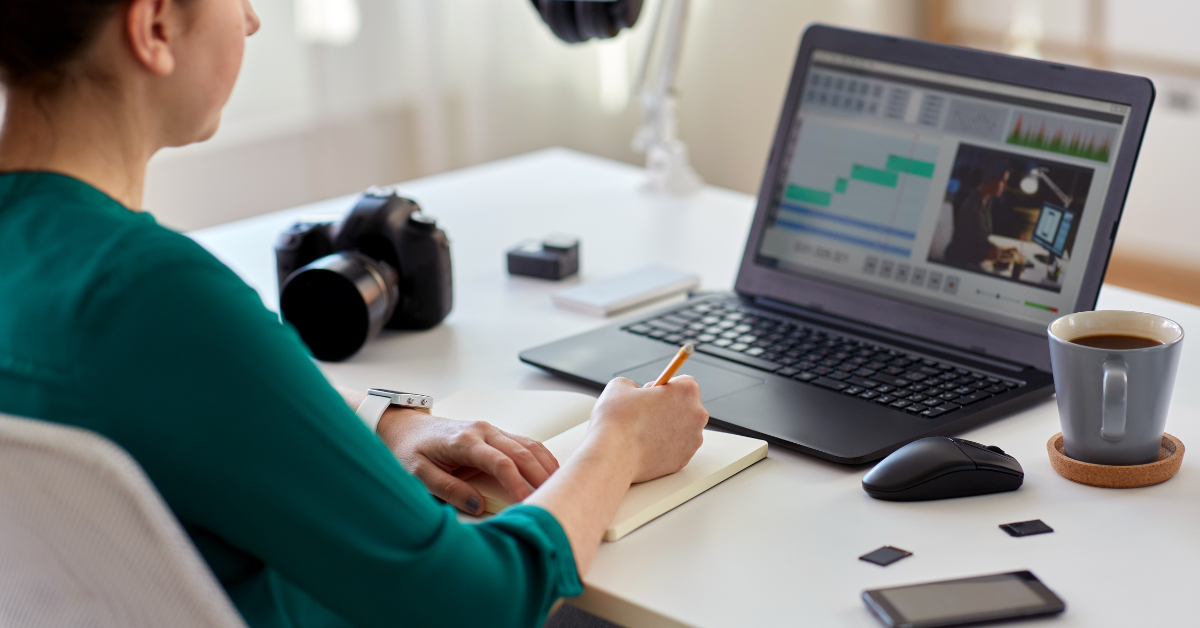 A person in a green shirt sits at a desk, taking notes in a notebook while looking at a laptop displaying video editing software. Engrossed in video content creation, they're surrounded by a camera and lens, memory cards, and a cup of coffee.