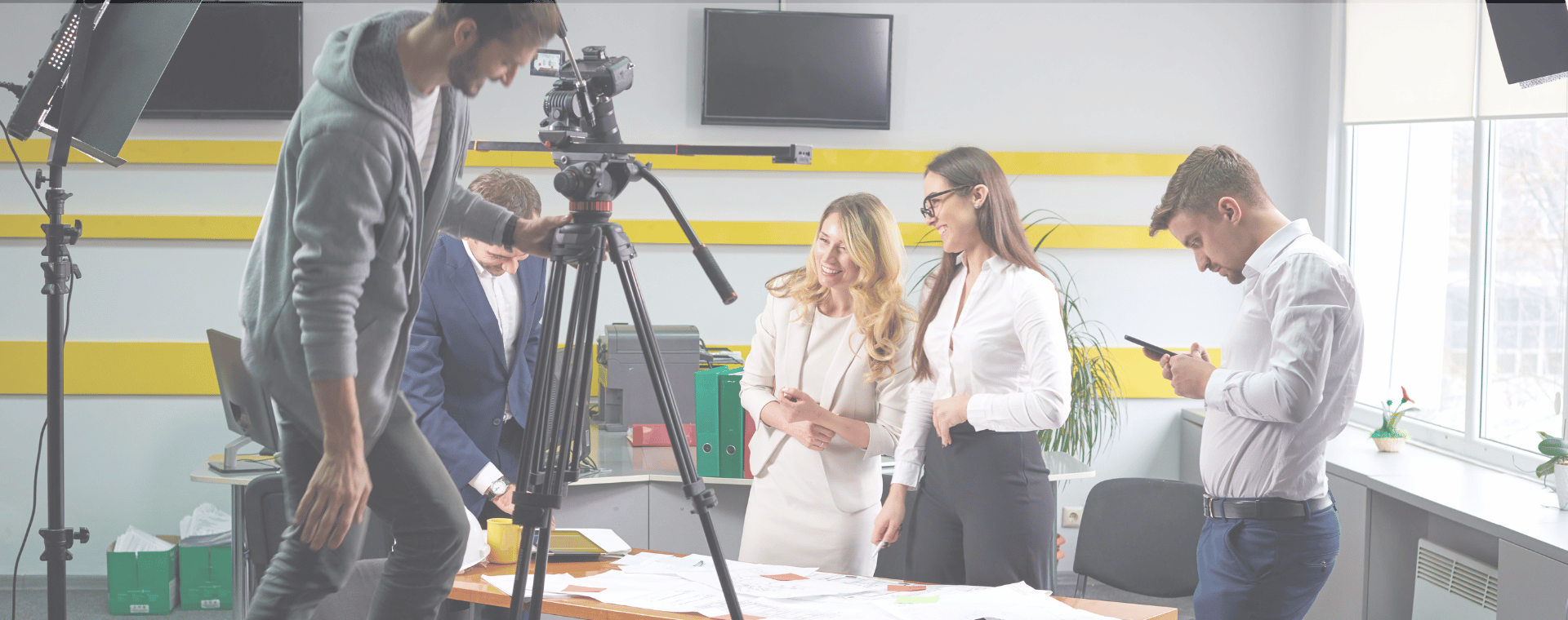 Four people are engaged on the set of a video shoot in a modern office. One man operates a camera on a tripod, another reads a script, and two women stand together, reviewing documents spread on a table. Video production equipment and monitors are visible around them as they focus on producing video content.