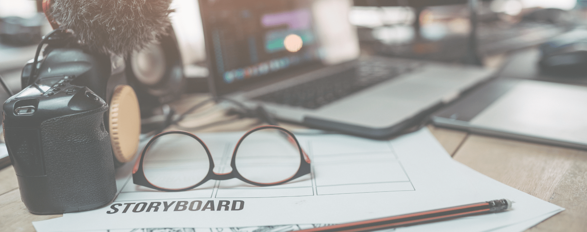 A pair of glasses rests on a storyboard sheet next to a pencil. In the background, there is a camera with a microphone attached, and an open laptop displaying video editing software. The scene depicts the animation production process in a workspace for video creation.