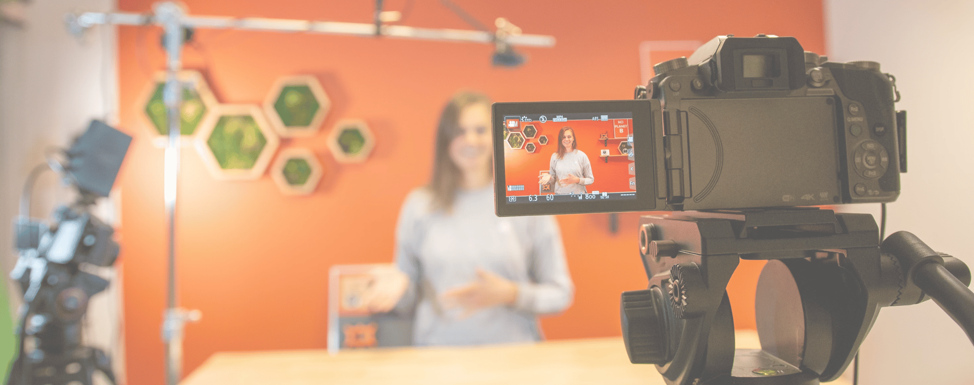 A marketing manager stands in front of an orange backdrop with hexagonal wall art, speaking to the camera. Multiple cameras and lighting equipment from a video production company are set up around her, capturing the scene. The focus is on the camera screen displaying her image.